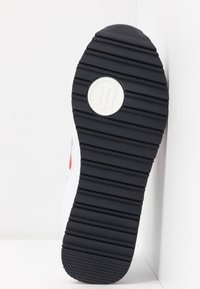 Semelle de chaussure noire avec des rainures horizontales et un logo circulaire blanc présentant des lettres entrelacées, attachée à une chaussure blanche et rouge.