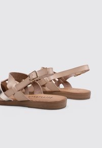 Sandalias de cuero beige con diseño entrecruzado y un acento metálico. Presentan una suela tostada y una correa ajustable con hebilla. Plantillas texturizadas visibles.