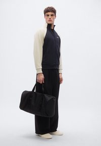 Laivastonsininen ja kerma vaatteissa, jossa on korkea kaulus ja vetoketju, yhdistetty mustiin leveälahkeisiin housuihin ja mustaan duffel-laukkuun. Beige-liukukengät.