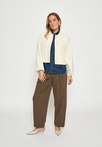Witte cropped jas over een denim shirt, gecombineerd met bruine wijde broek en beige puntige schoenen. Zachte texturen en knoopdetails.