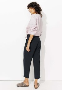 Femme aux cheveux bouclés portant une chemise rayée rose clair, un pantalon noir coupé, et des chaussures à enfiler imprimé léopard, se tenant contre un mur blanc.