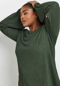 Top verde testurizzato con maniche lunghe a sbuffo, caratterizzato da un collo drappeggiato e un motivo di pieghe verticali. Tessuto morbido con una leggera lucentezza.