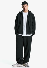 Zwarte zip-up hoodie met een koordje, gecombineerd met zwarte sweatpants. De outfit heeft een witte t-shirt eronder en grijze sportschoenen.