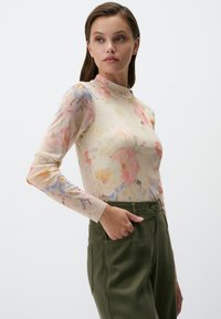 Top floreale a maniche lunghe in tessuto trasparente con colletto alto, con fiori rosa, blu e gialli su sfondo crema, abbinato a pantaloni verdi.