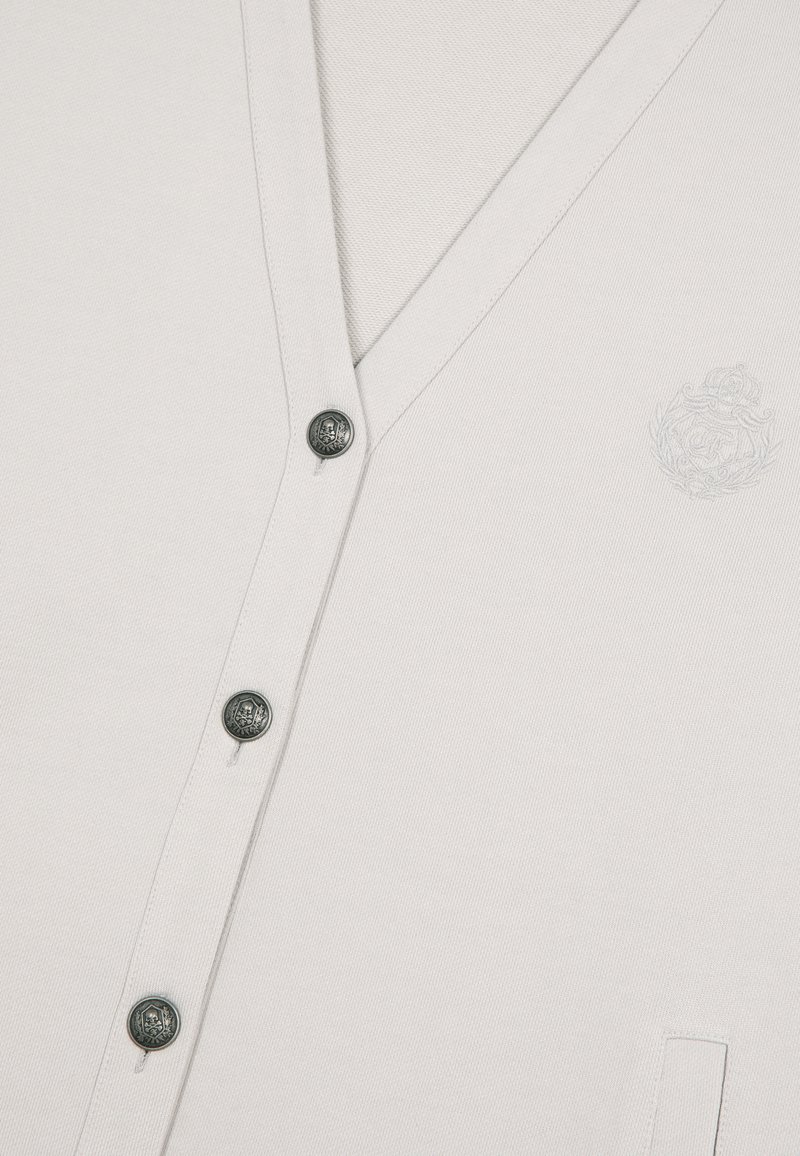 Weißer V-Ausschnitt-Cardigan mit Metallknöpfen und dezent gesticktem Wappen auf der Brust, das Textur und Nähdetails des Fabrics zeigt.