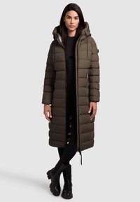 Lang olivengrønt dunjakke med hætte, der har et vandret quiltet design, frontlynlås og sidelommer, parret med sorte ankelstøvler.