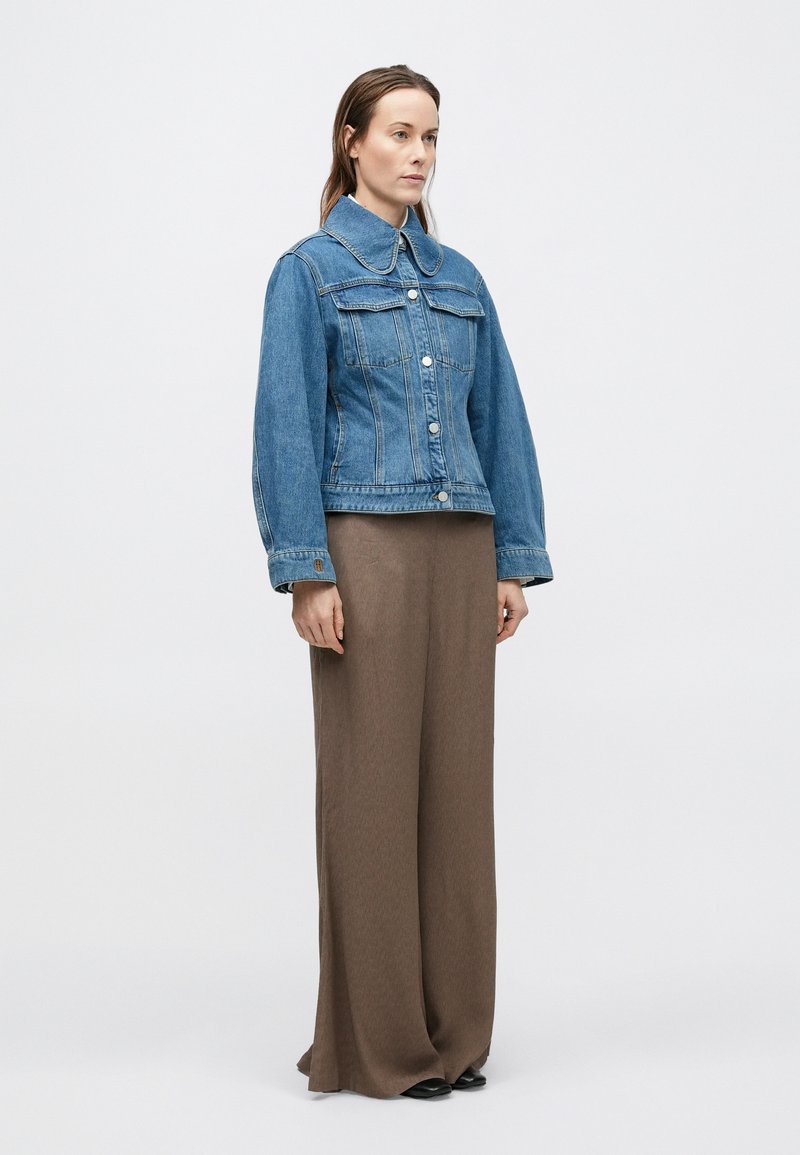 Denim jacket in medium blue with a structured collar, front button closure, and two chest pockets. Paired with long brown pants.