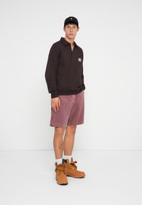 Young man wearing a black long-sleeve polo, pink shorts, tan work boots, white socks, and a black baseball cap, standing against a white background.