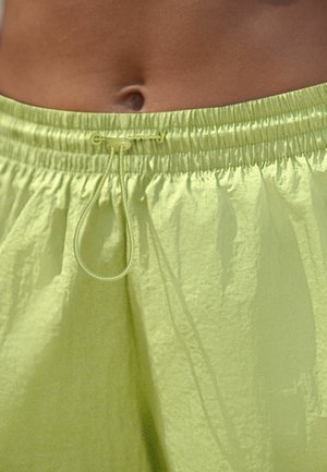 Pantaloni scurți verzi deschis strălucitori cu șnur și talie elastică purtați de o persoană care arată abdomenul și buricul.