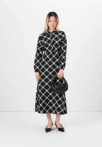Femme se tenant dans une longue robe à motif losange noir et blanc, tenant un sac à main noir, portant des talons aiguille noirs, devant un fond blanc uni.
