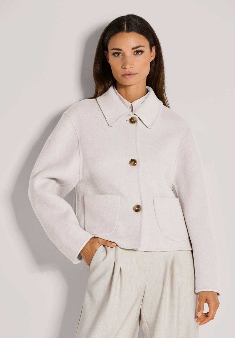 Vrouw in een lichtbeige cropped jas met grote knopen, een brede kraag en twee voorzakken, gecombineerd met losse crèmekleurige broeken.