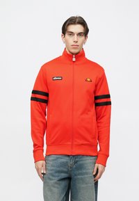 Punane tõmblukuga dressipluus kõrge kraega, mustade triipudega varrukatel, rinnal logomärk, sile tekstuur ja küljetaskud.