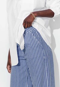 Chemise blanche à manches retroussées, rentrée dans un pantalon large rayé bleu et blanc. Mise en avant d'un gros plan d'une main reposant dans la poche.