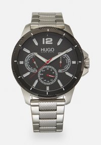 Orologio Hugo Boss con un quadrante nero texturizzato, dettagli argentati, lancetta dei secondi rossa e un cinturino in acciaio inossidabile a rete. Include visualizzazioni della data e del giorno.