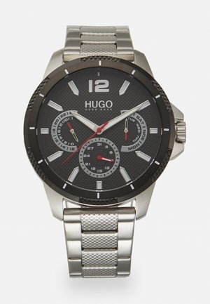 Reloj Hugo Boss con un dial texturizado negro, acentos plateados, segundero rojo y una correa de malla de acero inoxidable. Presenta visualización de fecha y día.