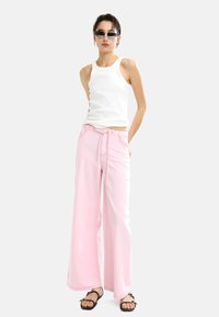 Biały top na ramiączkach, różowe szerokie spodnie z troczkiem w pasie i czarne sandały. Błyszcząca tekstura i kontrastowe szwy na spodniach.