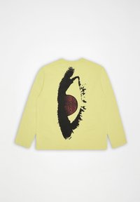 Κίτρινο πουκάμισο με μακριά μανίκια, με μεγάλο μαύρο και κόκκινο αφηρημένο σχέδιο ματιού τυπωμένο στην πλάτη, σε απλό λευκό φόντο.