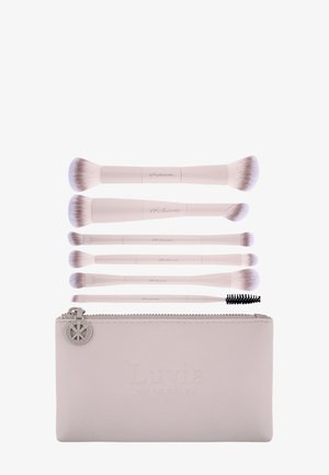 Ensemble de six pinceaux de maquillage à double embout avec manches blancs et poils doux, présentés au-dessus d'une pochette à pinceaux beige à fermeture éclair.