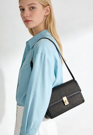 Jonge vrouw met lang blond haar draagt een lichtblauw knoopblouse, witte broek en draagt een getextureerde zwarte schoudertas met een gouden sluiting.