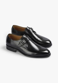 Zapatos de cuero negro con acabado pulido, punta puntiaguda y una correa decorativa con hebilla en la parte superior. Forro interior suave y tacón sólido.