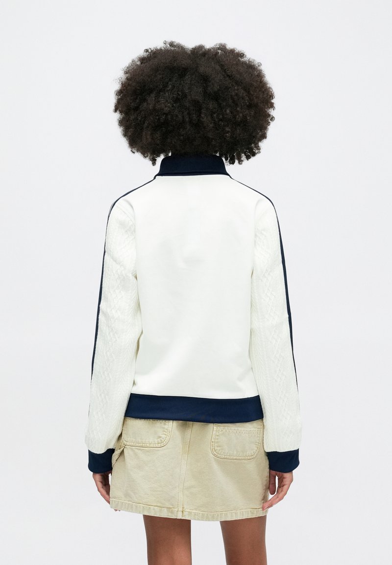 White and navy bomber jacket with knitted sleeve texture, high collar, and ribbed cuffs, paired with a beige skirt.