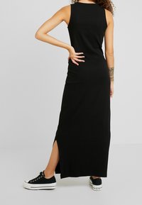 Person med lockigt hår som bär en ärmlös svart maxiklänning med ett sidoklyv, svart och vita sneakers, och en mönstrad tatuering på underarmen, stående.