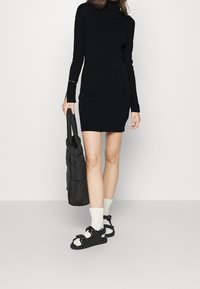 Robe en maille noire à manches longues et col montant. Associée à des sandales noires matelassées et des chaussettes courtes gris clair, tenant un sac noir.