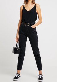 Schwarzes ärmelloses Oberteil mit V-Ausschnitt, hoch-taillierte schwarze Jeans, einen breiten Gürtel mit goldener Schnalle und schwarze Sneaker. Hält eine kleine schwarze Tasche.