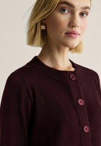 Vinröd cardigan med rund halsringning, tre knappar, mjuk textur och ribbade ärmslut. Designen är enkel och figurnära.