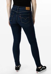 Jeans ajustados de denim azul oscuro con cinco bolsillos y una etiqueta de cuero marrón en la cintura. Combinados con zapatillas negras.