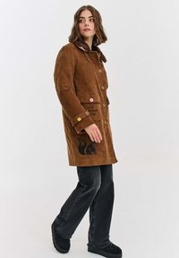 Brązowy płaszcz z korduroyu z kołnierzem z sztucznego futra, z przodu z guzikami, kieszeniami po bokach i czarnym wzorem lisa na dole. W zestawieniu z ciemnymi dżinsami.