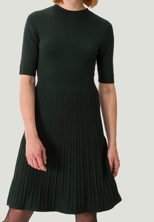 Femme portant une robe en maille côtelée vert foncé avec des manches courtes et une taille cintrée sur un fond clair uni.