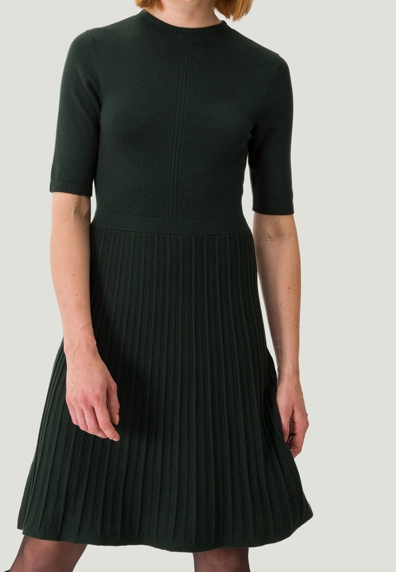 Femme portant une robe en maille côtelée vert foncé avec des manches courtes et une taille cintrée sur un fond clair uni.