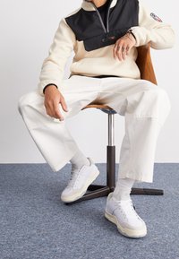 Vita och grå sneakers med texturerade detaljer, kombinerat med lösa vita byxor och en krämfärgad fleece-tröja med svart dragkedja.