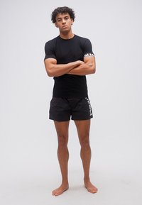 Jeune homme aux cheveux bouclés debout pieds nus, les bras croisés, portant une chemise noire à manches courtes et un short noir avec du texte blanc sur les manches et la jambe.