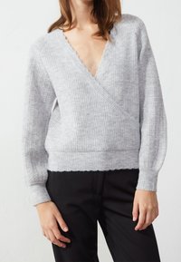 Light gray knitted sweater with a deep V-neck, ribbed texture, wide sleeves, and a fitted hem, paired with black trousers.