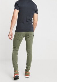 Olivgröna slim-fit jeans med detaljer på bakfickorna, tillsammans med en mörk t-shirt och sneakers med orange accenter. Slät tygstruktur.