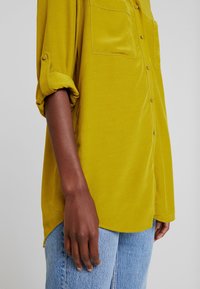 Senapsgul knapp-up skjorta med två bröstfickor, uppvikta ärmar och en mjuk textur, matchad med ljusblå jeans.