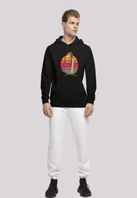 Felpa nera con cappuccio e una grafica colorata di un personaggio in un design circolare al tramonto, abbinata a pantaloni jogger bianchi e scarpe da ginnastica nere.