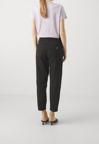 Pantalones negros de talle alto con perneras ajustadas, terminados con una pequeña etiqueta rectangular de la marca en el bolsillo trasero. Camiseta de cuello redondo en un tono lila claro.