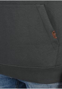 Sudadera gris oscura con dobladillo y puños acanalados, con un bolsillo frontal y una pequeña etiqueta de marca marrón en un lado. Textura suave.