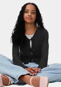 Zwarte, gestructureerde cardigan met een sluiting aan de voorkant, gedragen over een lichtgrijze top, gecombineerd met lichtblauwe wijde jeans en roze schoenen.