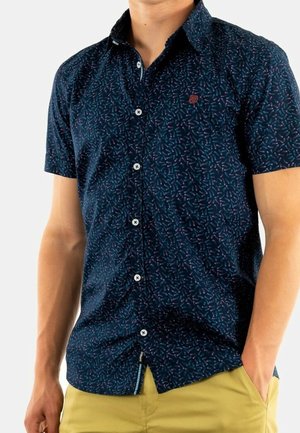 Homme portant une chemise bleu marine à manches courtes avec un petit motif abstrait et un pantalon jaune, les mains dans les poches.