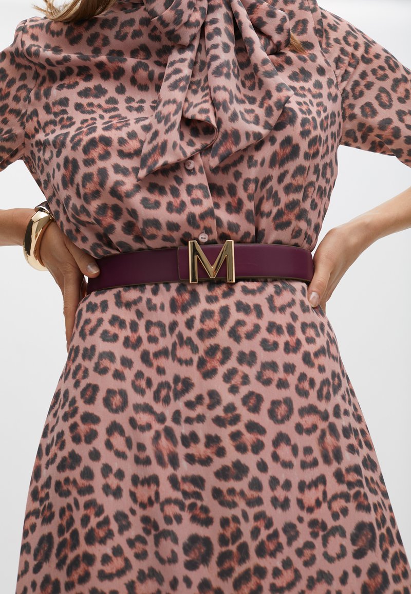 Halenka s leopardím vzorem a výstřihem s mašlí, nošená s širokým bordó koženým pásem s zlatou sponou ve tvaru písmene 'M'. Texturovaná a vzorovaná látka.
