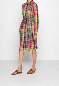 Frau in einem bunten karierten Hemdblusenkleid mit Gürtel und langen Ärmeln, die mit braunen flachen Sandalen vor einem schlichten weißen Hintergrund steht.