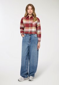 Camicia a quadri rossa, beige e bianca con bottoni e maniche lunghe; abbinata a jeans in denim azzurro chiaro, larghi, e sneakers argentate.