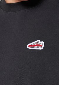 Camisa negra con cuello redondo, que presenta un parche bordado blanco de una zapatilla en rojo y gris con detalles en negro.