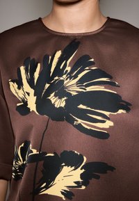 Blusa marrone con ampia stampa floreale nera e gialla, tessuto liscio, scollo rotondo e maniche corte.