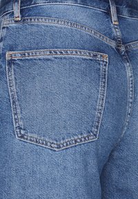 Jean en denim bleu avec une poche arrière en forme triangulaire et des coutures orange contrastantes. La texture semble lisse avec un léger motif de tissage.