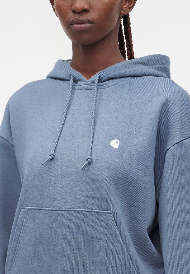 Woman wearing a blue hooded sweatshirt with drawstrings and a small white logo on the chest, braided hair visible on one side.